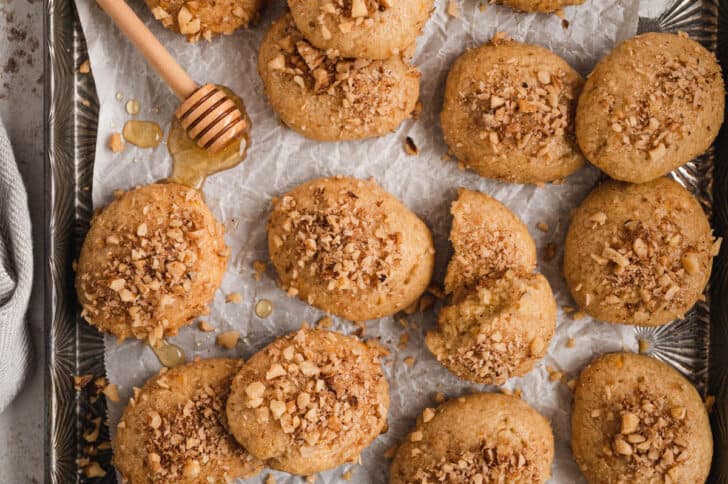 Melomakarona cookies sprinkled with walnuts on white parchment paper with a honey dipper.