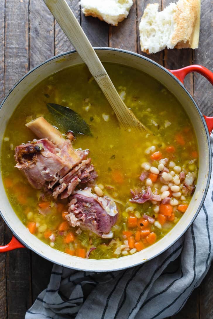 Ham bone soup with beans simmering in a red dutch oven with a wooden spoon stirring it.
