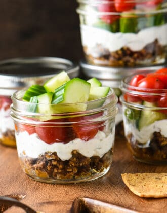 Pint sized mason jars filled with Greek layer dip, including ground lamb, yogurt, cucumbers and tomatoes.