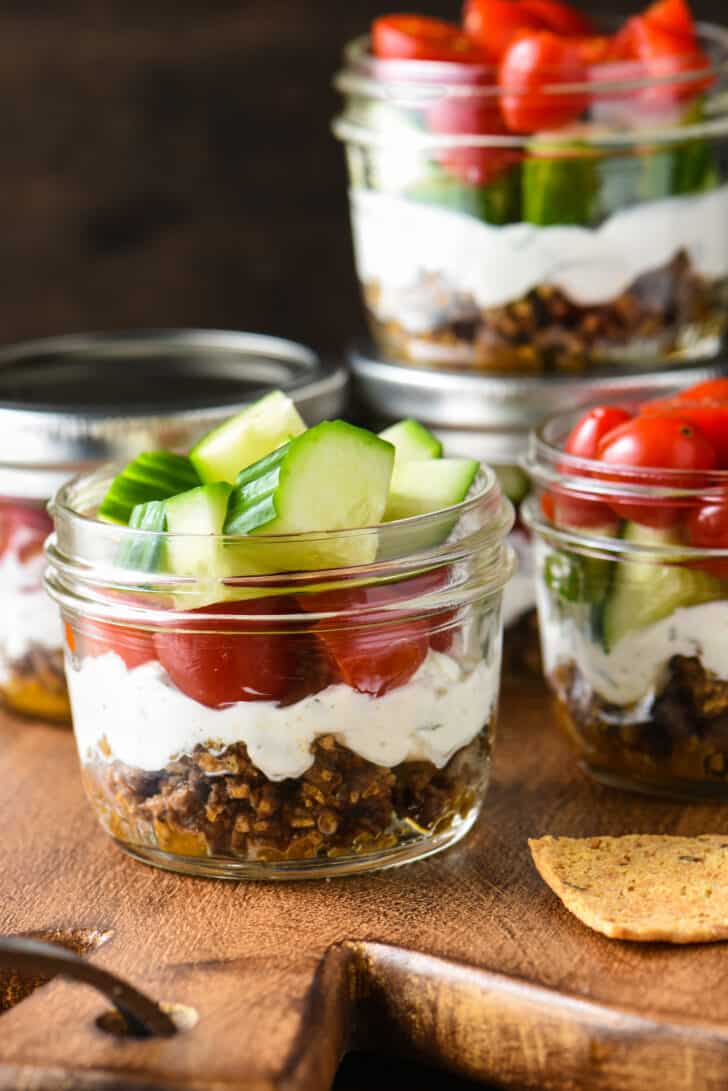 Pint sized mason jars filled with Greek layer dip, including ground lamb, yogurt, cucumbers and tomatoes.