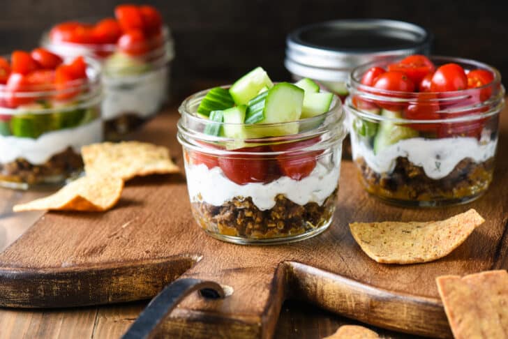 Pint sized mason jars filled with a layered Greek appetizer dip, including ground lamb, yogurt, cucumbers and tomatoes.