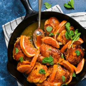 Cast iron skillet filled with orange glazed chicken thighs sprinkled with mint, with spoon scooping out sauce.