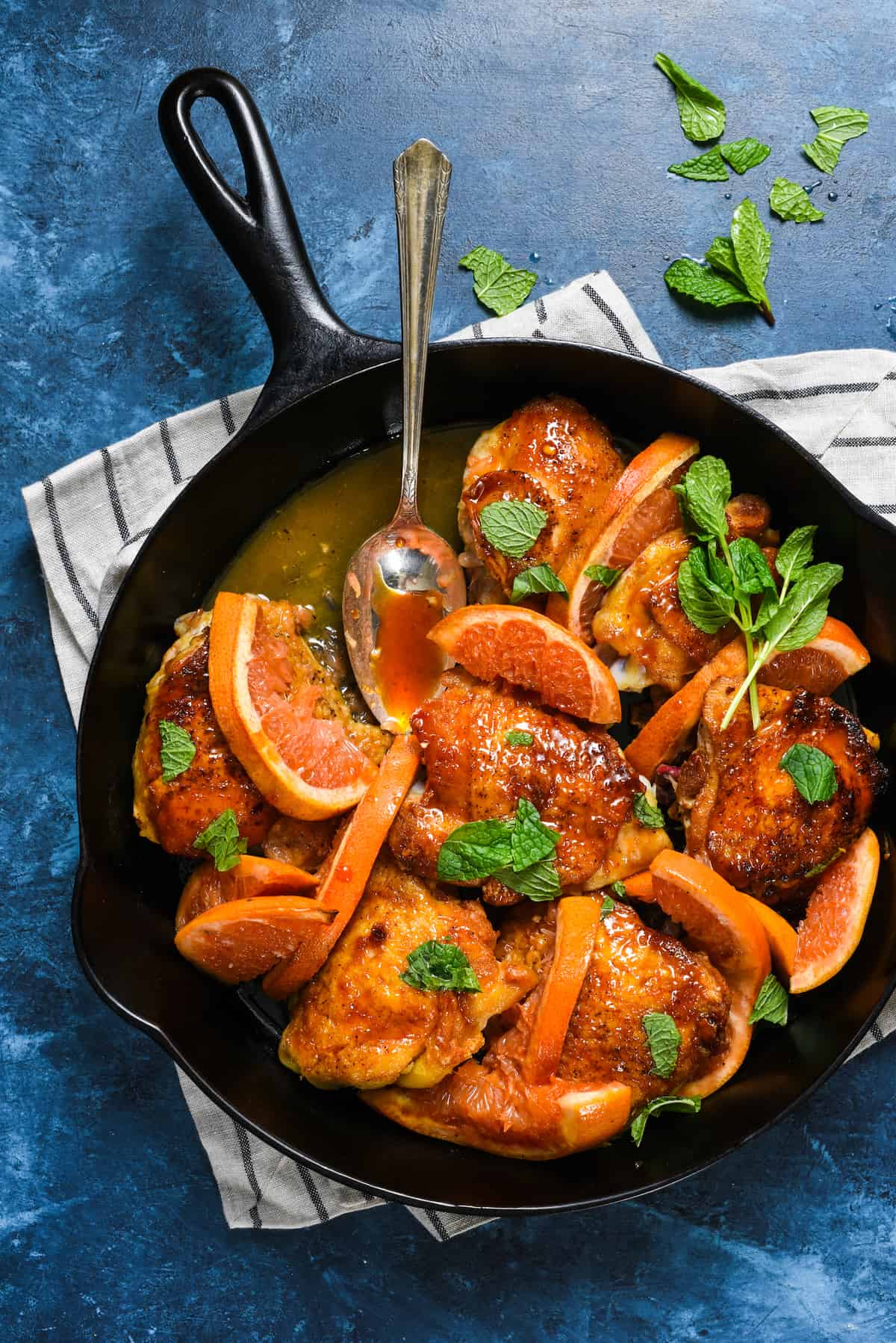 Cast iron skillet filled with orange glazed chicken thighs sprinkled with mint, with spoon scooping out sauce.