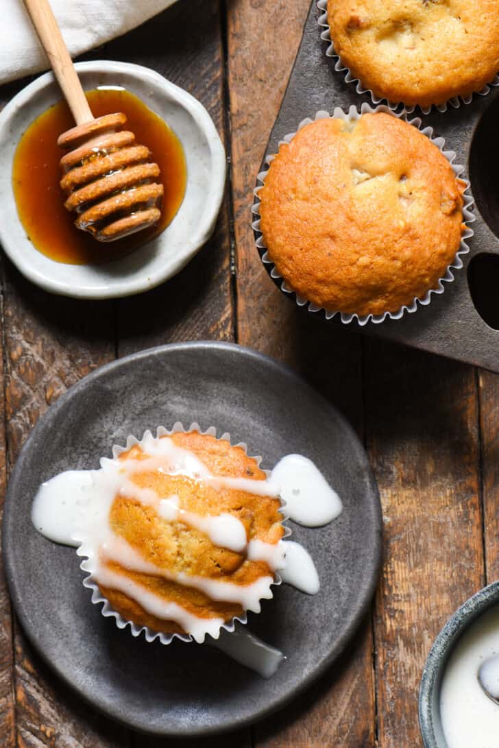 A small gray plate topped with a baked good in a cupcake wrapper, drizzled with glaze, alongside a small dish of honey.