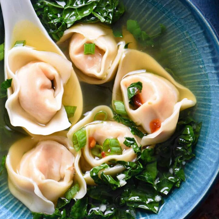 Shrimp wonton soup with kale in a blue bowl with a white soup spoon scooping up one of five wontons.