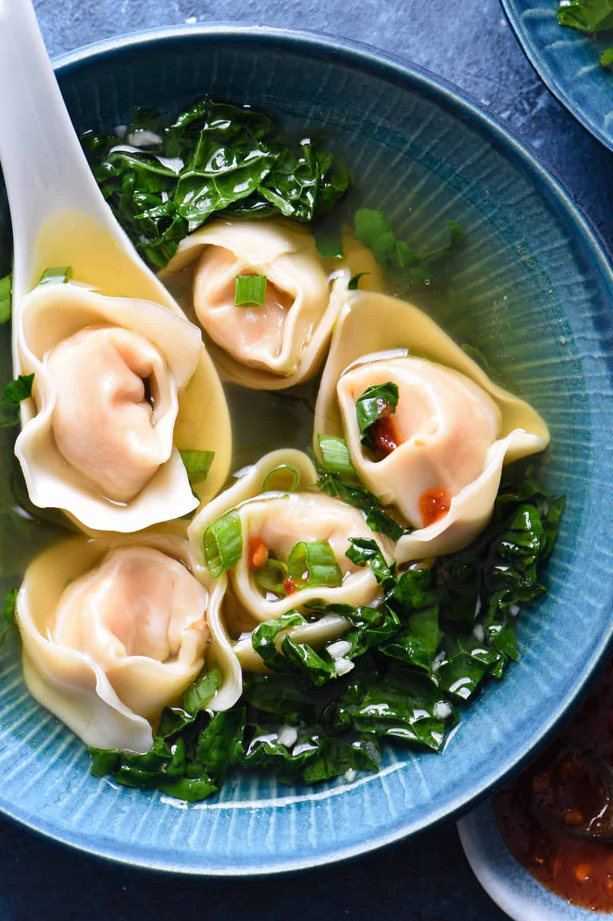 Shrimp wonton soup with kale in a blue bowl with a white soup spoon scooping up one of five wontons.