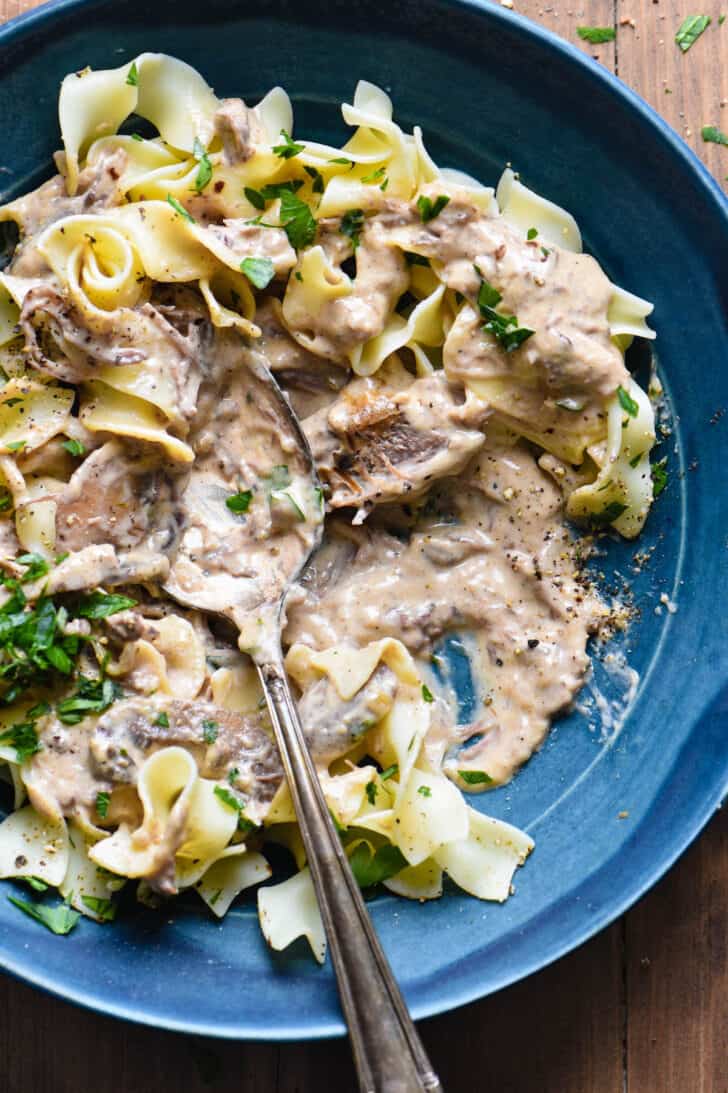 Slow cooker beef stroganoff over egg noodles, served in a blue bowl with a spoon scooping a bite.
