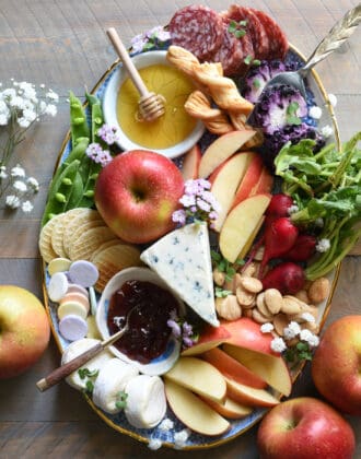 Oval platter filled with cheeses, fruit, honey, jam, nuts, meats and flowers.