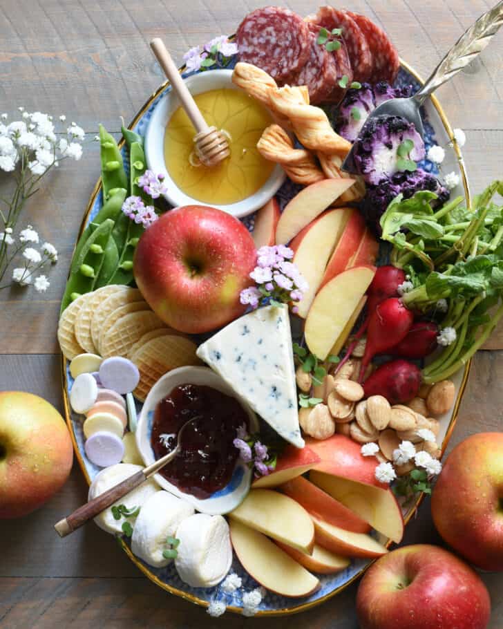 Oval platter filled with charcuterie board ideas like cheeses, fruit, honey, jam, nuts, meats and flowers.