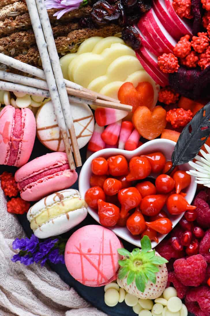 A platter of heart shaped food including cheese and bell peppers, along with crackers, radishes, candy, macarons and white chocolate.