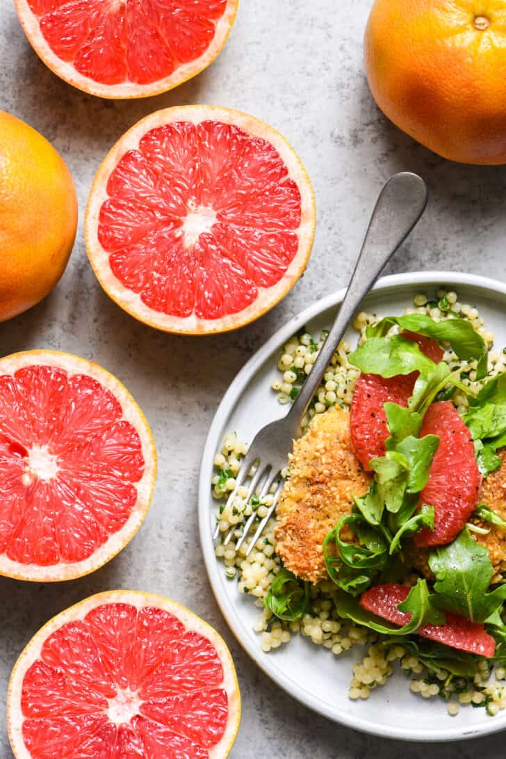 Four grapefruit halves alongside a plate filled with almond crusted chicken, couscous and salad.