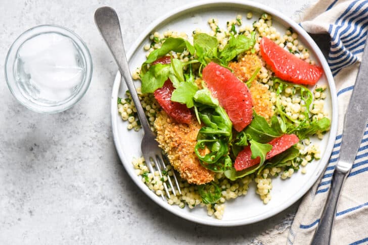 A small white plate of baked almond chicken with couscous, salad and grapefruit on a gray surface with a water glass nearby.