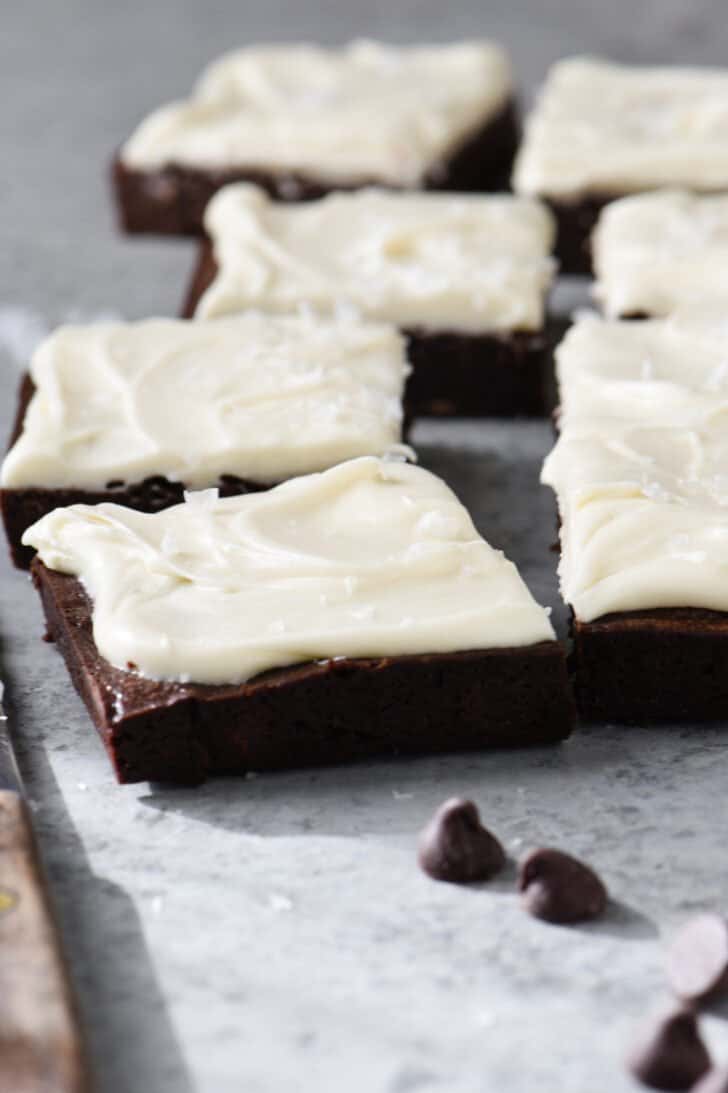 Chocolate Baileys brownies with cream cheese frosting, cut into squares, on a gray table with chocolate chips garnishing the scene.