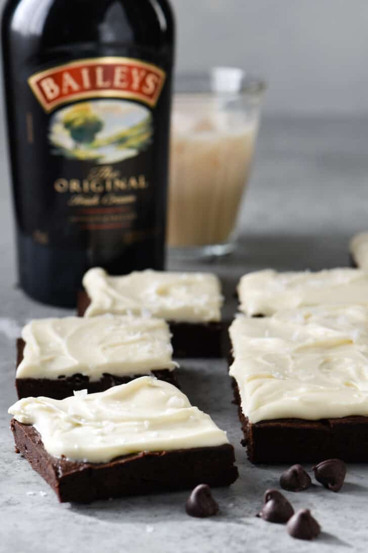 Irish cream brownies topped with fluffy white frosting, on a gray table, with a bottle of Baileys Irish Cream in the background.