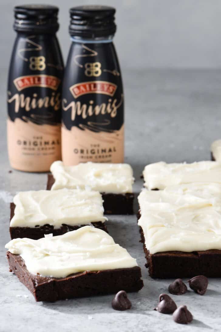 Irish cream brownies topped with fluffy white frosting, on a gray table, with two small bottles of Baileys Irish Cream in the background.