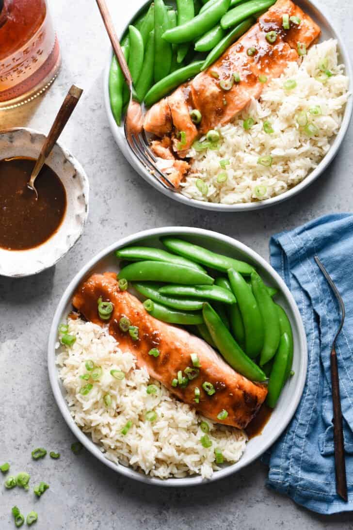 Two gray plates filled with bourbon glazed salmon and white rice garnished with green onions, as well as sugar snap peas.