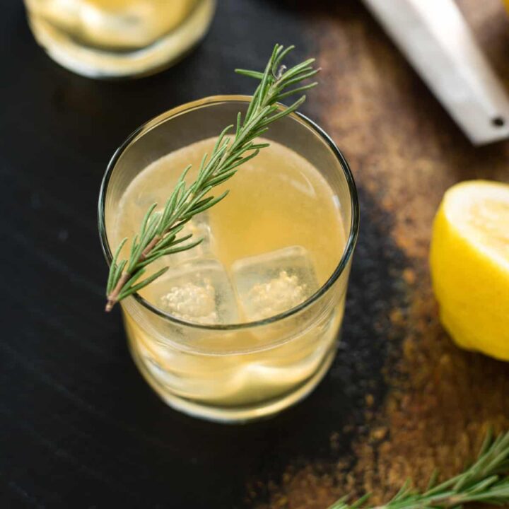 A lemon hued cocktail with a sprig of rosemary garnishing the glass.