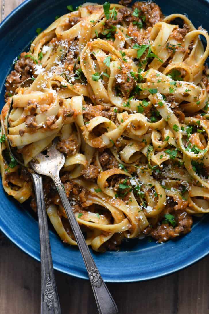 A blue ceramic bowl filled with fettuccine bolognese, with a fork twirling the pasta.