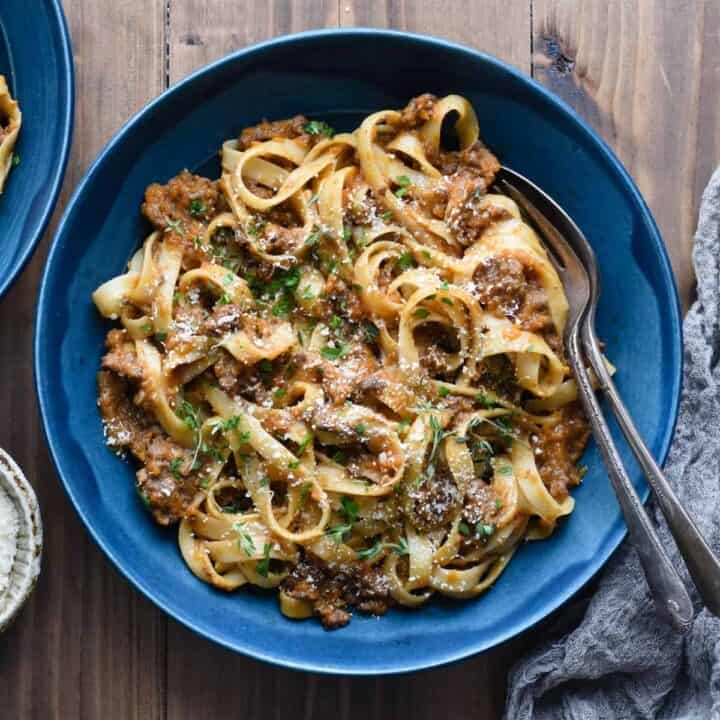 A blue ceramic bowl filled with fettuccine bolognese, with a fork and spoon resting in the bowl.