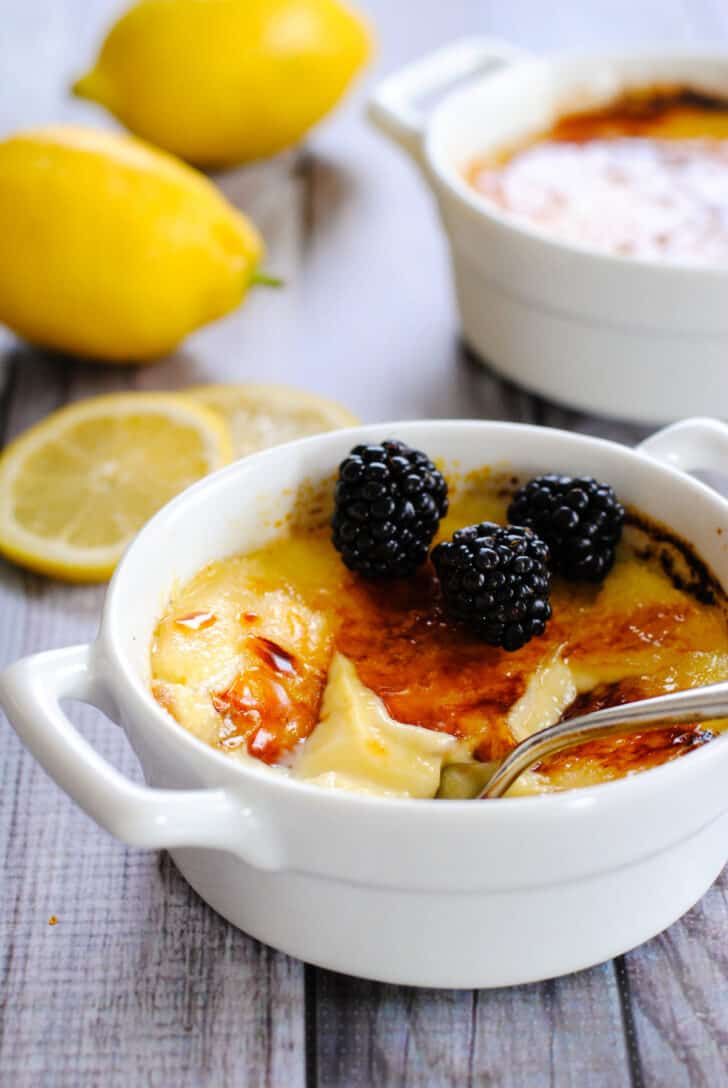 A dessert scene of creme brulee for two, with two white ceramic bowls with handles, filled with lemon creme brulee and topped with blackberries.