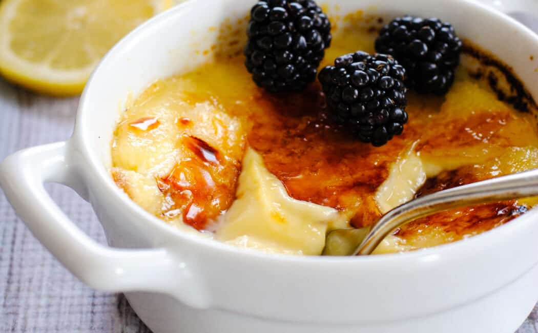 White ceramic bowl with handles, filled with lemon creme brulee and topped with blackberries.