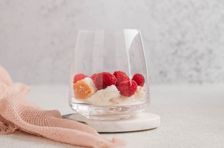 A stemless wine glass with layers of cubed angel food cake and raspberries in the bottom.