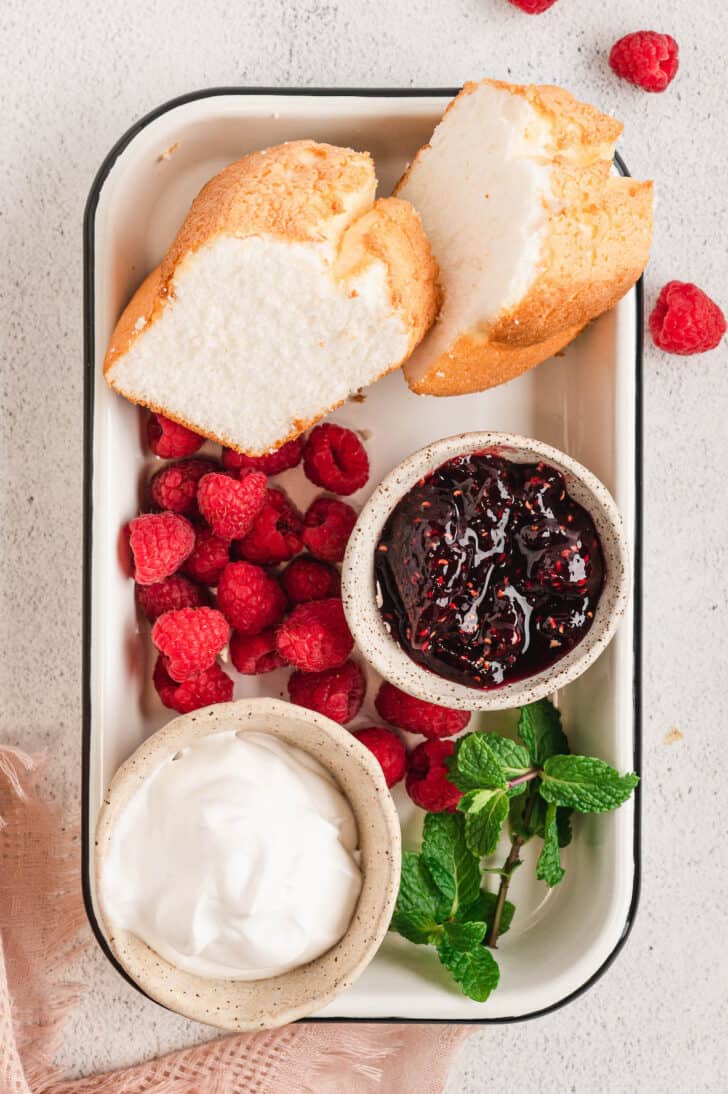Ingredients needed for a Valentines dessert, on a tray, including raspberry jam, fresh raspberries, angel food cake, whipped cream, and fresh mint.