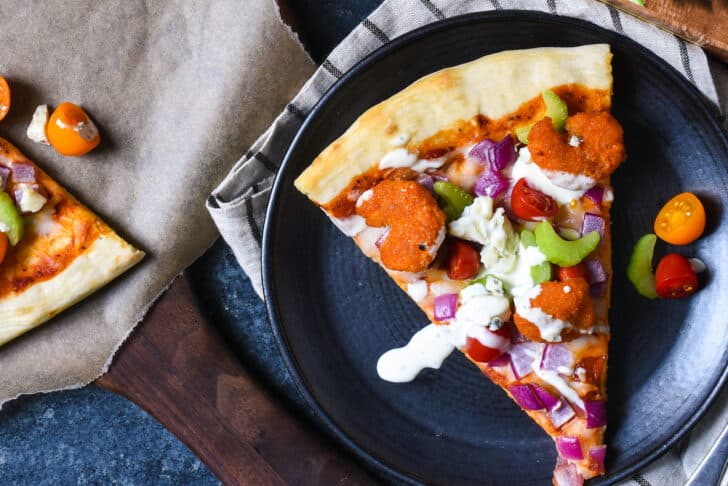 A slice of flatbread on a black plate,  topped with vegetables, spicy seafood, cheese and ranch dressing.