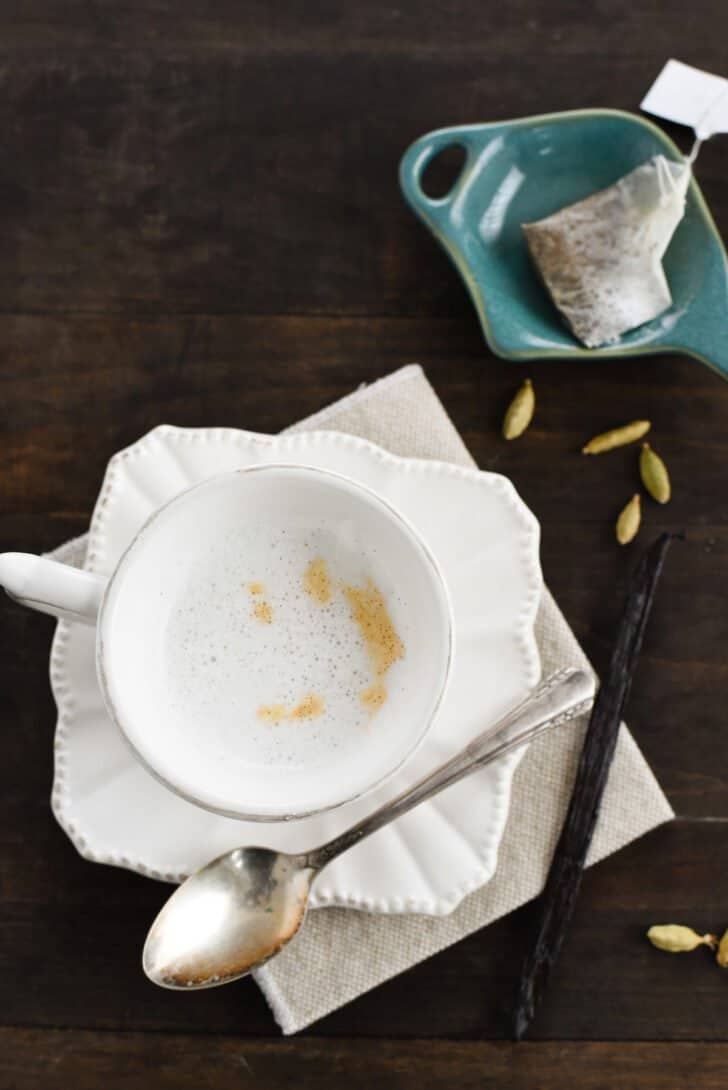 A small white teacup filled with frothy cardamom tea on a matching saucer, with a spoon alongside.