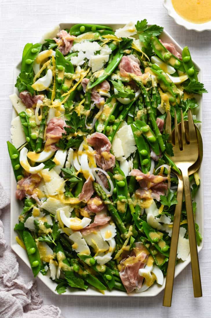Large white platter filled with spring salad made with asparagus, snap peas, prosciutto, cheese and shallots.