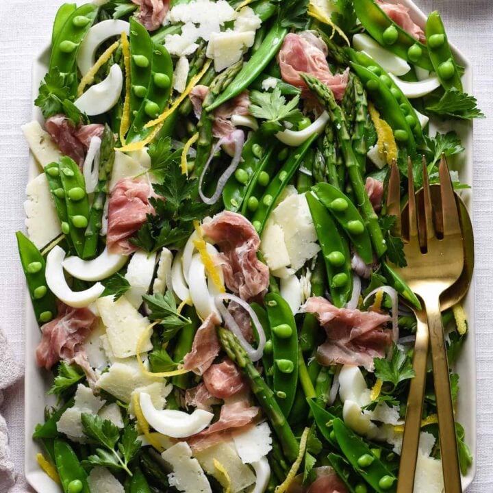 White platter filled with spring salad made with asparagus, snap peas, prosciutto, cheese and shallots.