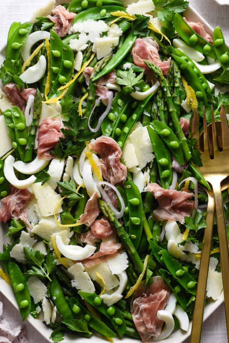 Closeup on a large platter of spring salad made with peas, asparagus, shallots, hard boiled eggs, cheese and cured meat.