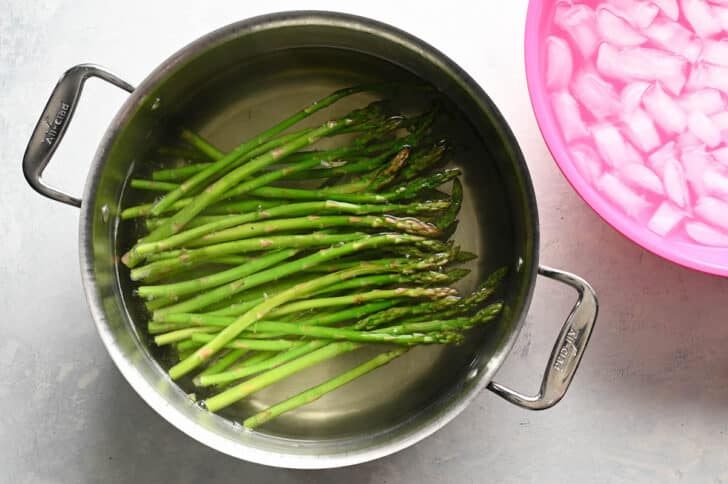 A large stainless steel pot filled with water and asparagus, with a pink bowl of ice water next to it.