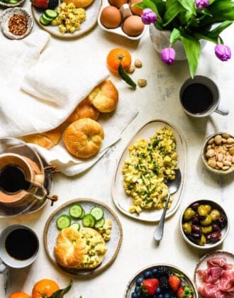 A large brunch buffet set up on a white tabletop, including pickled egg salad, croissants, fruit, olives and flowers.