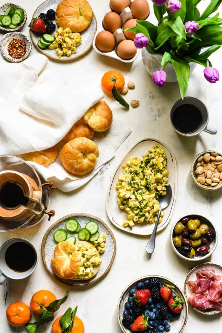 A large brunch buffet set up on a white tabletop, including pickled egg salad, croissants, fruit, olives and flowers.