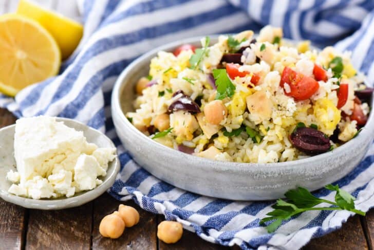A gray bowl sits on top of a blue and white striped napkin, filled with Mediterranean fried rice made with chicken, tomatoes, olives, herbs and feta cheese.