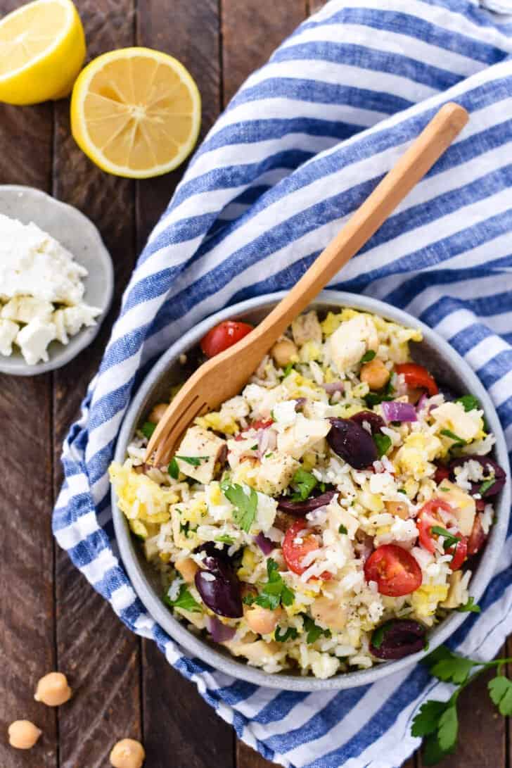 A gray bowl sits on top of a blue and white striped napkin, filled with Greek fried rice made with chicken, tomatoes, olives, herbs and feta cheese.
