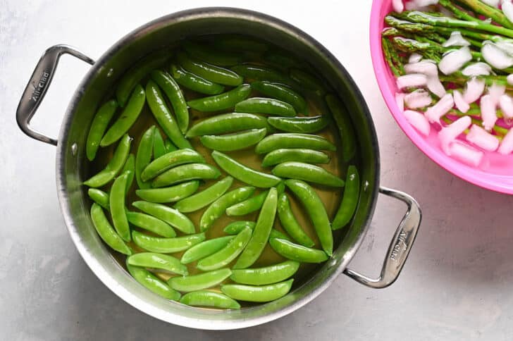 A large stainless steel pot filled with water and snap peas, with a pink bowl of ice water and asparagus next to it.