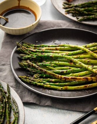 Roasted balsamic asparagus on a dark plate, with a small bowl of extra glaze in the background.