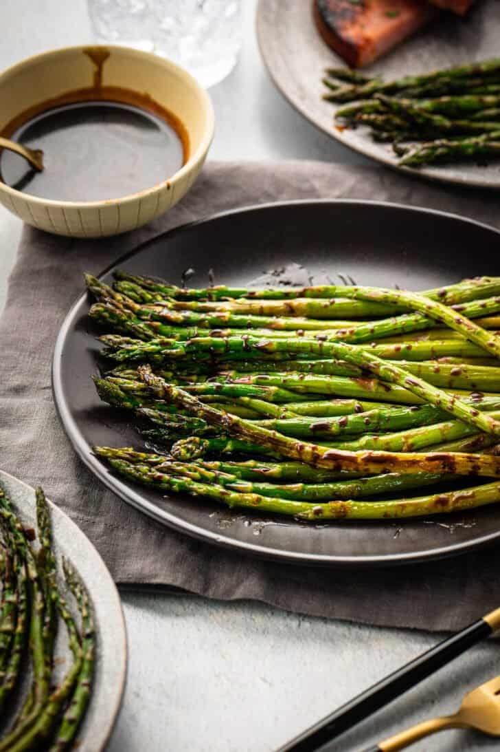 Roasted balsamic asparagus on a dark plate, with a small bowl of extra glaze in the background.