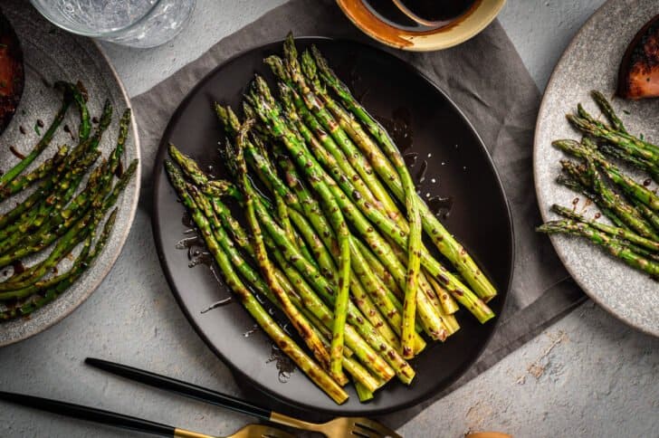 Asparagus with balsamic glaze on a dark platter, with dinner plates nearby.
