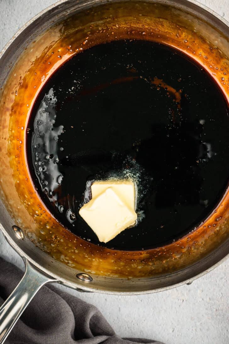 Two pats of butter being swirled into a skillet of dark liquid. 