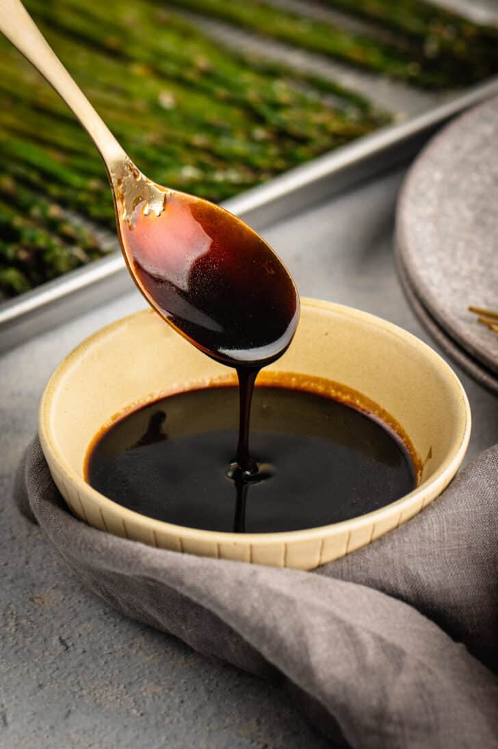 A gold spoon dripping balsamic glaze into a small beige bowl, with a tray of roasted asparagus in the background.