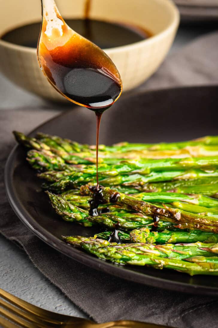 A gold spoon drizzling balsamic glaze over a bowl of roasted asparagus.