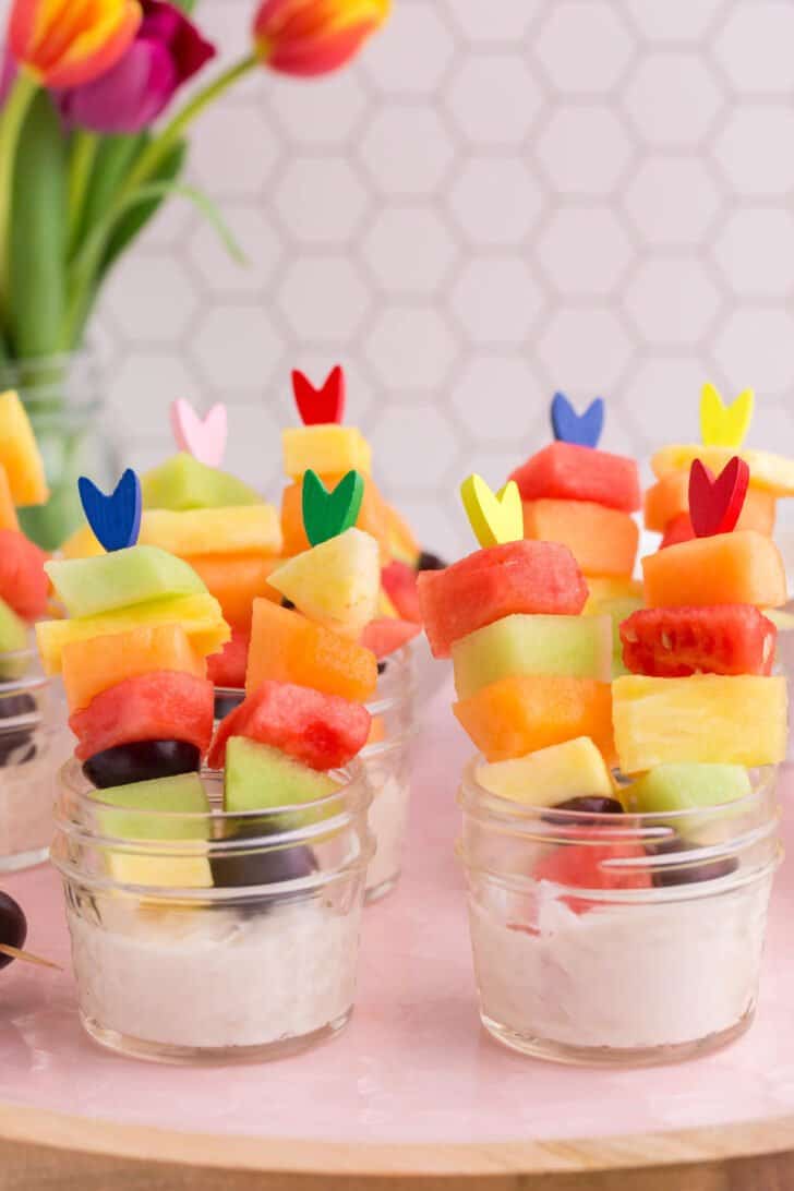 Small glass jars filled with white dip and skewers of fresh cut fruit.