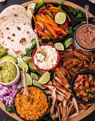 A large round wooden platter filled with fajita bar ingredients like tortillas, cooked chicken and steak, dips, cheese, rice, beans and lime.