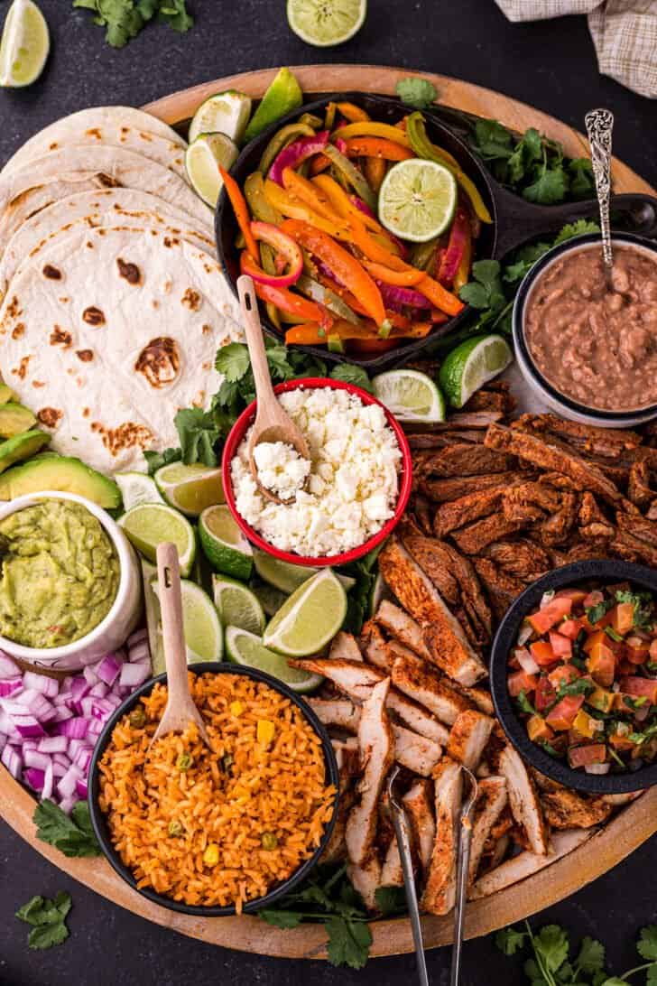 A large round wooden platter filled with fajita bar ingredients like tortillas, cooked chicken and steak, dips, cheese, rice, beans and lime.