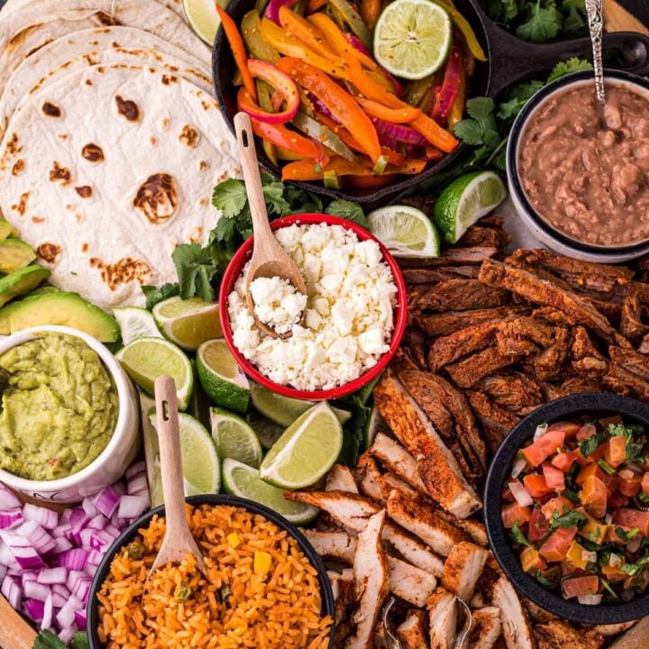 A large round wooden platter filled with fajita bar ingredients like tortillas, cooked chicken and steak, dips, cheese, rice, beans and lime.