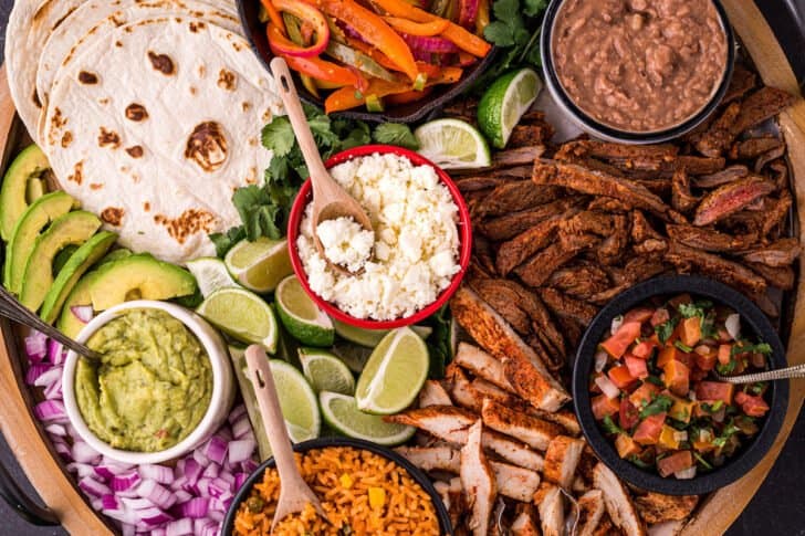 A large round wooden platter filled with fajita bar ingredients like tortillas, cooked chicken and steak, dips, cheese, rice, beans and lime.