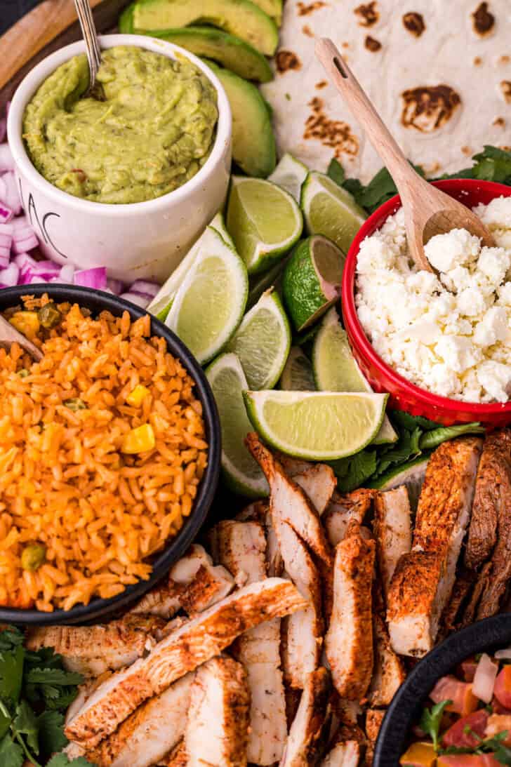 Mexican rice, cooked chicken, cotija cheese, lime wedges, guacamole, onions and tortillas arranged on a platter.